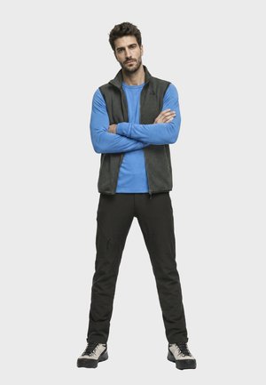 Mann mit Bart und kurzen lockigen Haaren steht mit verschränkten Armen, trägt ein blaues Langarmhemd, eine dunkelgraue Weste mit Reißverschluss, schwarze Hose und weiße Wanderschuhe.