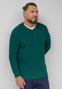 Zielona koszula z długim rękawem w stylu henley z teksturowanym wykończeniem, z trzema guzikami przy szyi i okrągłym kołnierzem, w połączeniu z ciemnymi spodniami.