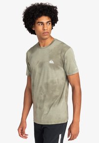 Camiseta de manga corta en verde olivo con cuello redondo, que presenta un sutil patrón de tie-dye y un pequeño logo blanco en la zona del pecho izquierdo.