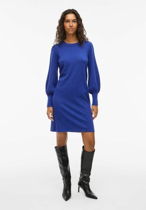 Femme aux cheveux bouclés portant une robe à manches longues bleu roi et des bottes noires montantes à talons sur fond blanc.