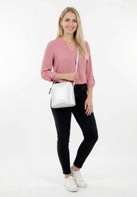 Young woman standing, wearing a pink blouse, black jeans, white sneakers, and a silver crossbody bag, smiling against a plain white background.