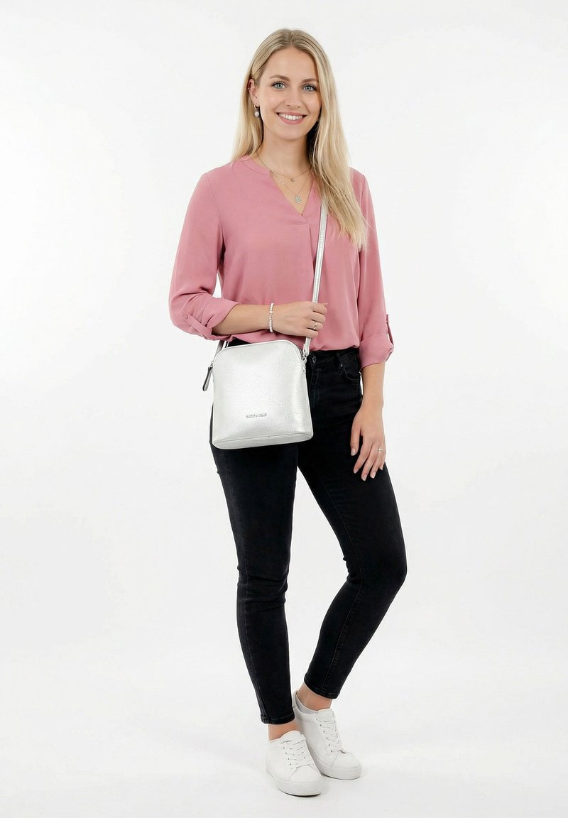 Young woman standing, wearing a pink blouse, black jeans, white sneakers, and a silver crossbody bag, smiling against a plain white background.