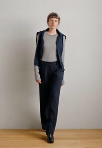 Camisola cinza de mangas compridas por baixo de um colete sem mangas azul escuro, combinada com calças azul-marinho com riscas e botas pretas até ao tornozelo.