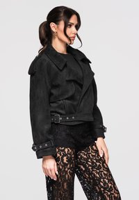 Femme portant une veste ceinturée en suède noir et un pantalon en dentelle noire, posant devant un fond gris clair uni.