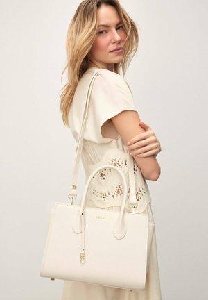 Donna con un vestito beige chiaro porta una grande borsa crema con due manici e una tracolla sulla spalla.