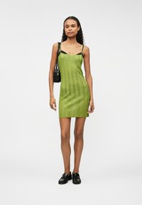 Zelené žebrované mini šaty s tenkými ramínky, spárované s malou černou kabelkou. Modelka má na sobě černou obuv a stojí na jednobarevném bílém pozadí.