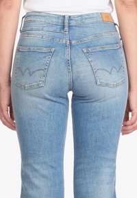 Hellblaue Jeans mit hoher Taille, auffälligen Gesäßtaschen mit Zickzack-Stichmuster und einem hellbraunen Leder-Patch am Bund.