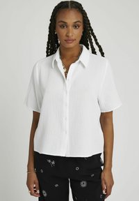 Chemise blanche à manches courtes avec boutons, en tissu texturé, présentant une coupe légèrement raccourcie et un col.