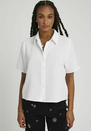 Chemise blanche à manches courtes avec boutons, en tissu texturé, présentant une coupe légèrement raccourcie et un col.