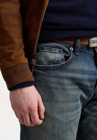 Muž v hnědé semišové bundě, námořnickém svetru, modrých džínách a hnědém koženém pásku s kovovou sponou, ruka uvolněně podél těla.