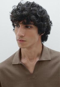 Polo a maniche corte, di colore marrone, con colletto a V. Il tessuto sembra morbido con una texture liscia. Il soggetto ha i capelli scuri e ricci, con uno sfondo neutro.