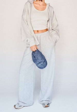 Person trägt hellgraue Jogginghose, passenden übergroßen Hoodie, weißes Croptop, hält blaue Jeanskappe, steht vor einfarbigem Hintergrund.