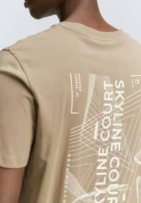 Pamučna t-shirt bež boje s kratkim rukavima, s bijelim grafičkim dizajnom koji uključuje tekst i geometrijske linije na leđima.