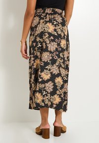 Jupe noire à motif floral avec une taille élastique, longueur mi-mollet, et un détail complexe beige et orange, associée à des sabots à talons couleur fauve.