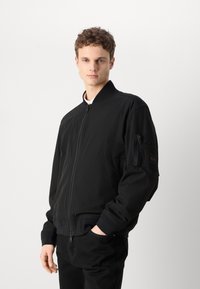 Černá bomber bunda se zipem vpředu a žebrovanými manžetami. Obsahuje boční kapsu na rukávu a má štíhlý design. Hladká texturovaná tkanina.