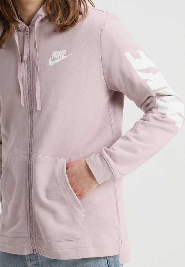 Sudadera con cremallera en color rosa, hecha de una tela suave. Presenta un logo de Nike en blanco en el pecho y texto en la manga izquierda. Incluye bolsillos frontales.