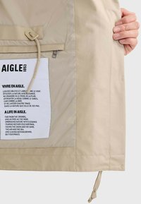 Beige, leichte Jacke mit Innenfutter, das ein Pflegeetikett mit schwarzem Text zeigt. An der Seite sind eine Kordelzugöffnung und eine Sichttasche mit Reißverschluss zu sehen.
