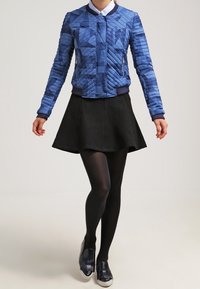 Blau gesteppte Bomberjacke mit geometrischen Mustern, geripptem Kragen und Reißverschlusstaschen, kombiniert mit einem schwarzen ausgestellten Rock und schwarzen Strumpfhosen.