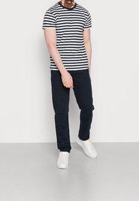 T-shirt w paski w granatowym i białym kolorze, krótki rękaw, okrągły dekolt; zestawiony z ciemnymi spodniami i białymi sneakersami, stojący na jasnej podłodze.