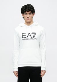 Mladý muž s kučeravými tmavými vlasmi, oblečený v bielej mikine Emporio Armani s čiernym logom a čiernych nohaviciach na jednoduchom bielom pozadí.