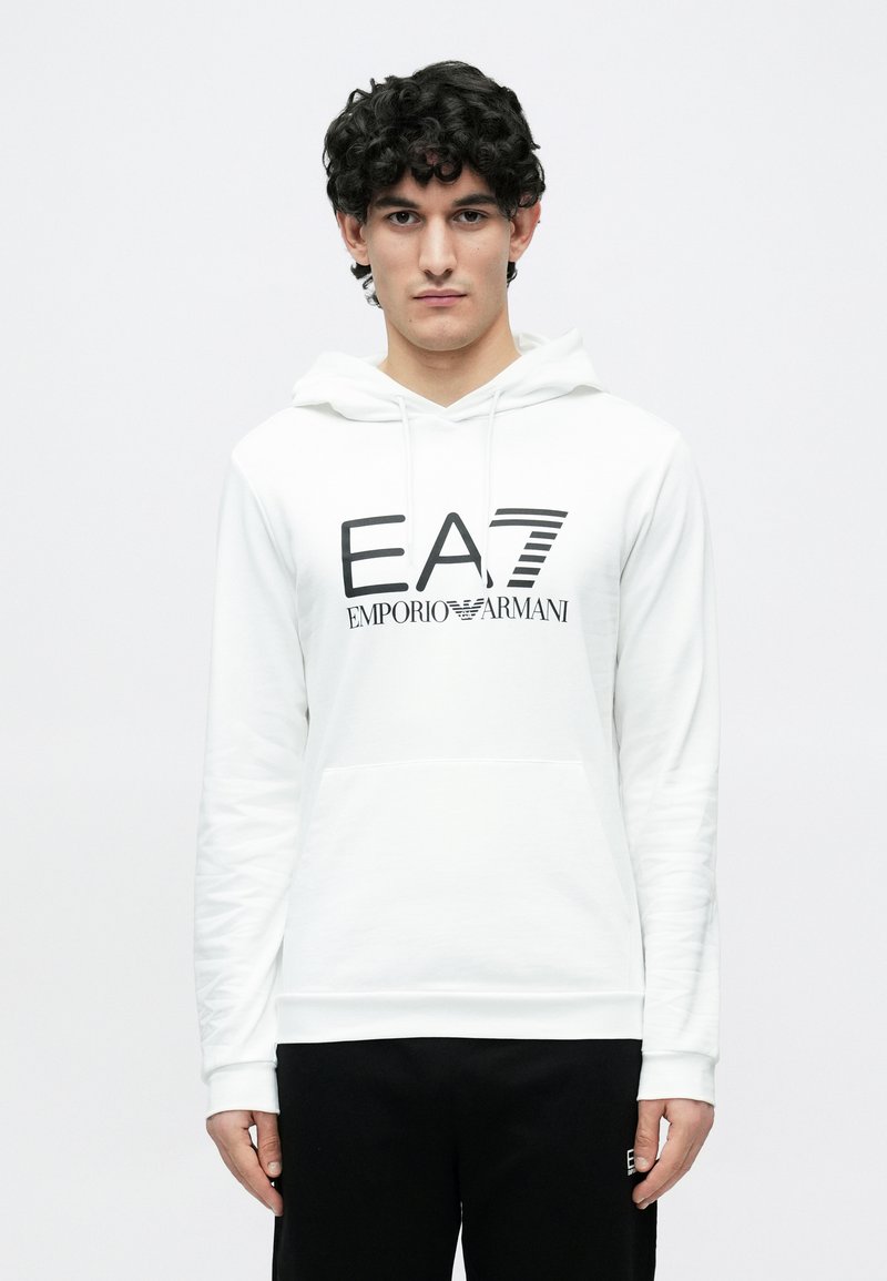 Mladý muž s kučeravými tmavými vlasmi, oblečený v bielej mikine Emporio Armani s čiernym logom a čiernych nohaviciach na jednoduchom bielom pozadí.