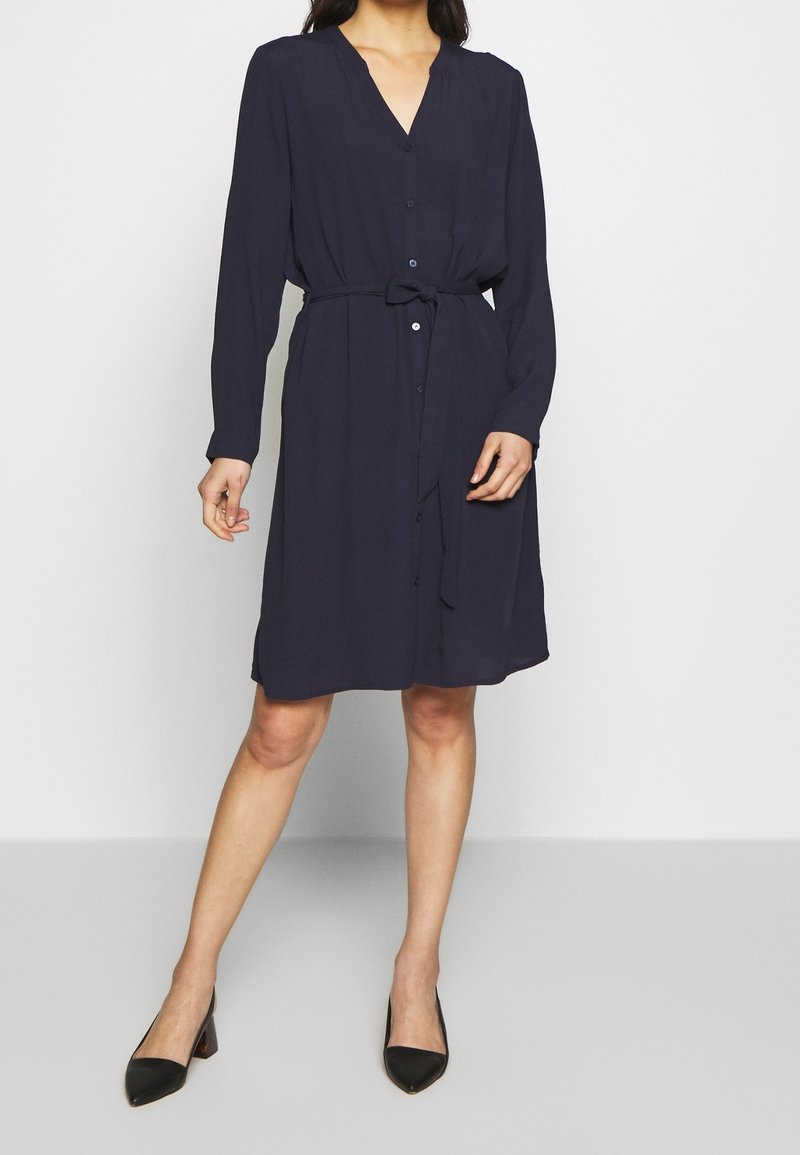 Femme portant une robe bleu marine à manches longues arrivant aux genoux, avec des boutons sur le devant, une ceinture à la taille et des escarpins noirs à bouts pointus, debout devant un fond uni.
