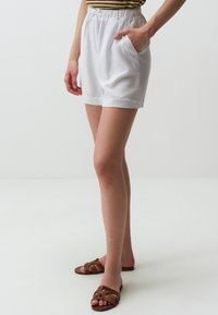 Pantaloni corti bianchi in lino con elastico in vita, dotati di tasche laterali. Abbinati a sandali a slide intrecciati marroni su uno sfondo neutro.