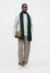 Pull vert clair avec motif texturé, écharpe bleu foncé, sac beige, pantalon marron et baskets blanches. Design simple et moderne.