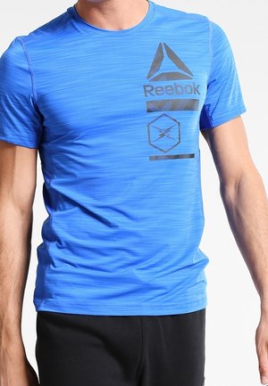 Uomo che indossa una maglietta blu aderente Reebok con loghi e pantaloni neri, mostrato dal collo ai fianchi su sfondo bianco.