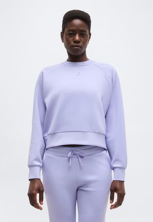 Persona che indossa una felpa a maniche lunghe di colore viola chiaro e pantaloni con coulisse abbinati, in piedi davanti a uno sfondo chiaro e uniforme.