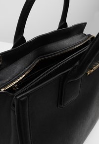 Musta käsilaukku, jossa on teksturoitu pinta, kultaiset vetoketjut ja useita osastoja. Rakenne on muotoiltu ja siinä on kaksi lyhyempää kahvaa.