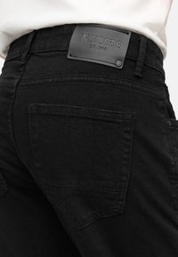Jeans de denim negro con una superficie texturizada, que presentan un parche de cuero etiquetado "INDICODE" en la cintura y detalles en el bolsillo trasero.