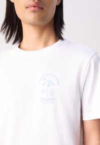 Bílý bavlněný t-shirt s jemným modrým grafickým potiskem, který obsahuje palmu, plážové prvky a text. Krátké rukávy a design s kulatým výstřihem.