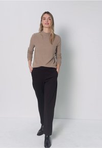 Pull côtelé marron clair à manches longues associé à un pantalon noir à jambes larges et des bottines noires, debout devant un fond neutre.
