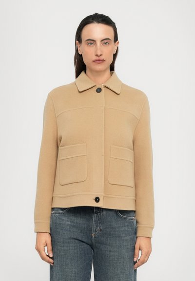 Mujer vistiendo una chaqueta de lana beige abotonada con bolsillos de solapa y pantalones vaqueros azules, de pie contra un fondo blanco liso.