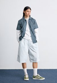 Kortermet stripete skjorte i blått og grønt, lagdelt over en hvit langermet skjorte; lyseblå shorts og grønne sneakers med hvite såler.