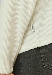 Top de tela blanca acanalada con una etiqueta en el dobladillo, combinado con unos jeans oscuros texturizados. El primer plano muestra el detalle de la manga y el contraste de materiales.
