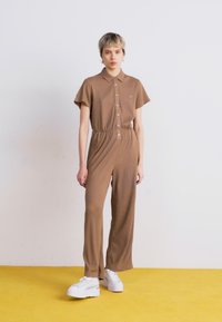 Jumpsuit marron à côtes avec des manches courtes et un col, doté d'une fermeture à boutons à l'avant et d'une taille élastique, associé à des chaussures à plateforme blanches.