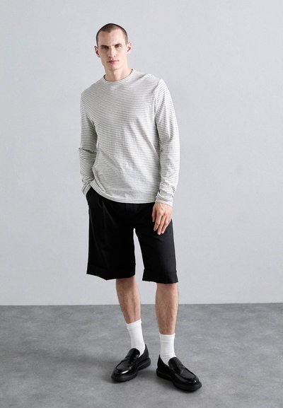 Camisa de mangas largas a rayas en gris claro y blanco, combinada con pantalones cortos negros, mocasines negros y calcetines blancos. Diseño de atuendo simple y casual.