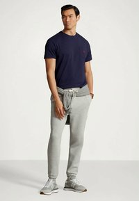 Mężczyzna w granatowej koszulce, szarych dresowych spodniach, szarych sneakersach i szarej bluzie przewiązanej w pasie, stojący na tle gładkiej ściany.