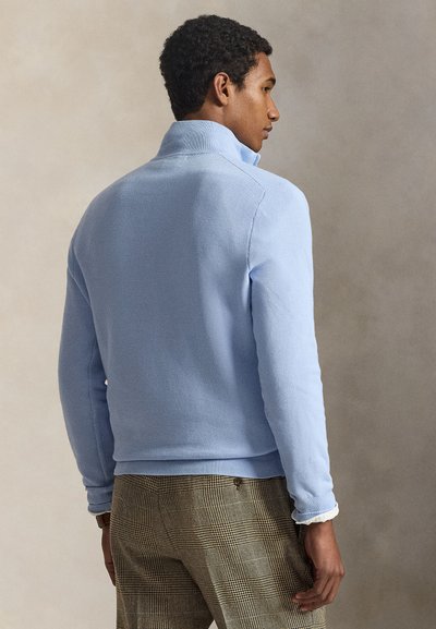 Mann in einem hellblauen Pullover mit hohem Kragen und braunen karierten Hosen, stehend und seitlich vor neutralem Hintergrund.