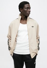 Bež trenirka jakna s crnim dizajnom tri pruge na rukavima, prednjim patentnim zatvaračem i izvezenim logotipom. Nošena preko bijele majice bez rukava i crnih hlača.