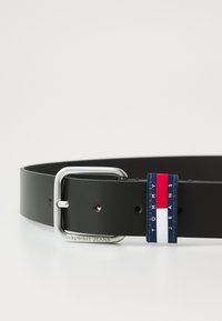 Zwarte leren riem met een zilveren metalen gesp en een blauwe, rode en witte logo-label met "TOMMY JEANS" erop gedrukt.