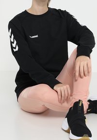 Sweat-shirt noir avec logo blanc, col rond large, et bandes chevron blanches contrastantes sur les manches, assorti à des leggings roses et des baskets noires.