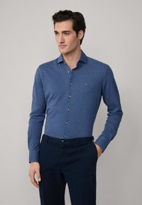 Hombre vistiendo una camisa de botones azul ajustada de manga larga y pantalones oscuros, de pie con una mano en el bolsillo frente a un fondo neutro.