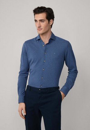 Hombre vistiendo una camisa de botones azul ajustada de manga larga y pantalones oscuros, de pie con una mano en el bolsillo frente a un fondo neutro.