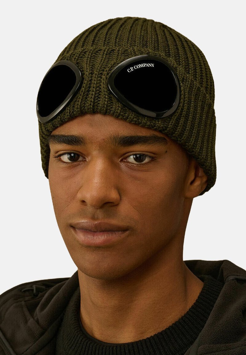 Beanie lavorato a maglia verde oliva con texture a coste, dotato di lenti nere a forma di occhiali integrate nel design, che offrono un accento unico.