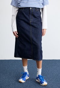 Gonna midi in denim blu scuro con cuciture gialle, abbinata a una camicia a righe blu e bianca e scarpe da ginnastica blu.