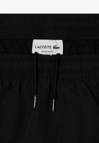 Juodi maudymosi šortai su tinklu, elastinga juosmens juosta ir raišteliu. Yra Lacoste etiketė ir sidabriniai logotipo akcentai ant raištelio.