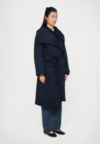 Manteau en laine bleu marine avec un large col, une ceinture et des poches ; présente une silhouette longue et sophistiquée ainsi qu'une texture lisse.
